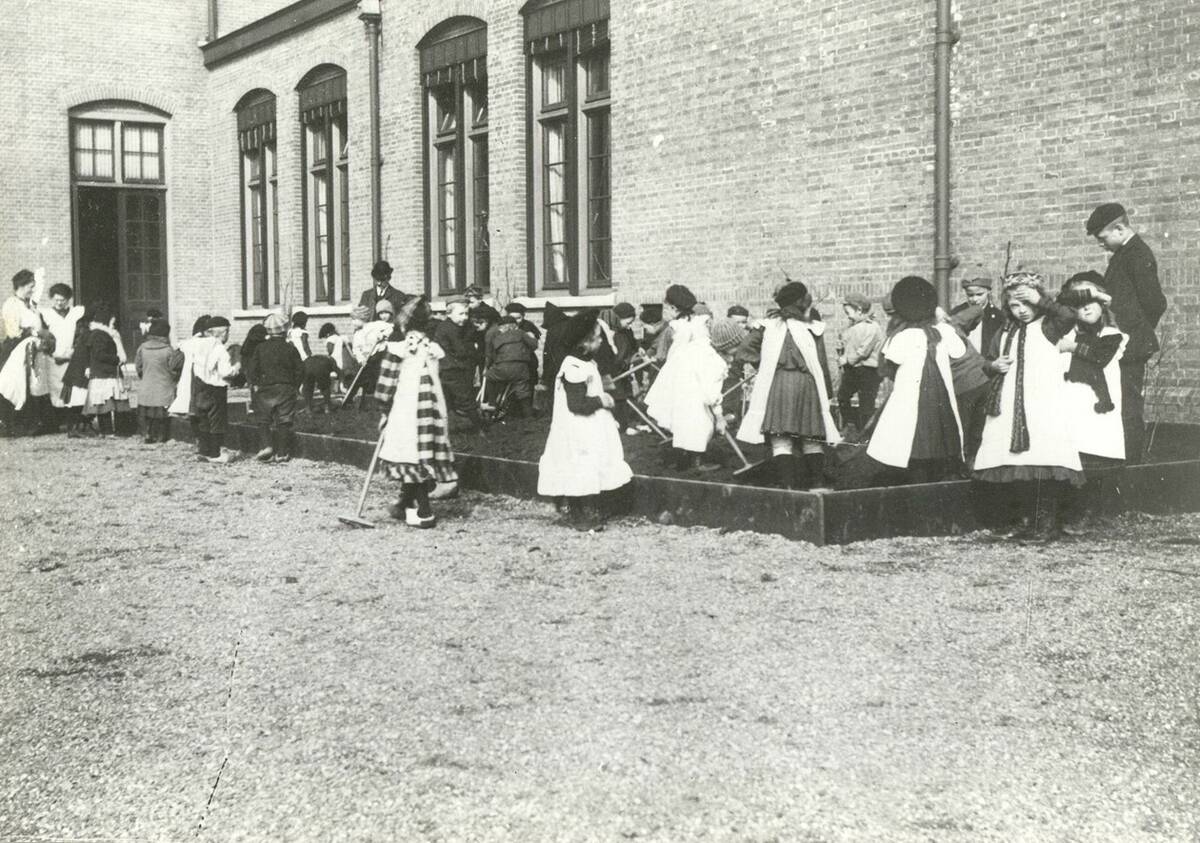 kindeen op klopmpen en laarzen, petten op en witte schorten om. Ze houden een harkje vast en staan in en om de tuinbakken met aarde