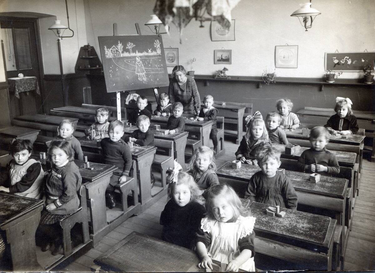 kinderen in houten schoolbankjes, met daarop voor elk een doosje blokjes. ze kijken in de camera maar lachen niet. De juf staat achterin en heeft een tekening op het bord gemaakt.